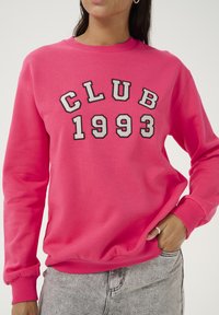 Rosa Sweatshirt mit dem Aufdruck "CLUB 1993" in fetten, kontrastierenden schwarz-weißen Buchstaben. Verfügt über lange Ärmel und gerippte Bündchen.