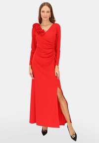 Czerwona sukienka z długim rękawem, z marszczeniem i kwiatowym akcentem na ramieniu; posiada boczne rozcięcie i gładką fakturę. Czarne, szpiczaste buty.
