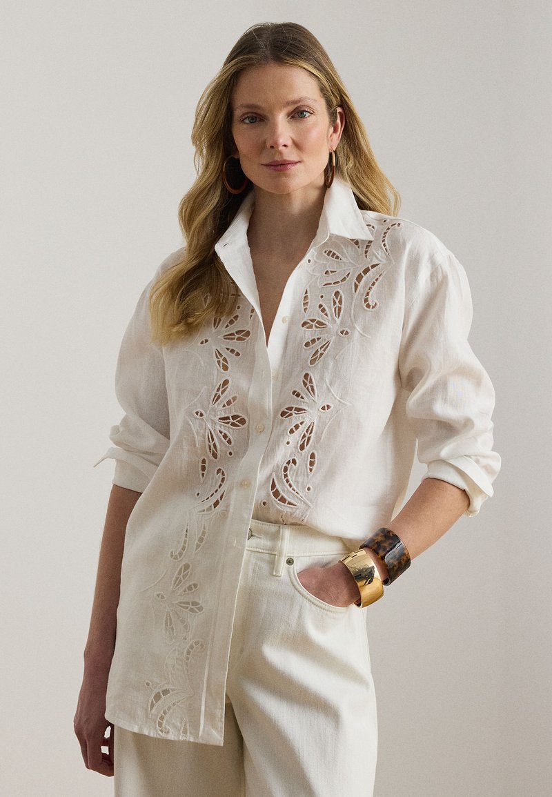 Nő fehér gombos ingben, melyen virágmintás kivágott hímzés díszíti, fehér nadrágban, és nagy arany- és teknőspáncél mintájú karkötőkben, zsebébe tett kézzel áll.