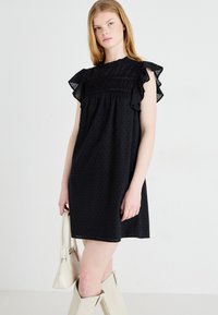 Μαύρο φόρεμα με ριγέ μανίκια και λεπτομέρεια φούσκας μπροστά. Φτιαγμένο από υφάσματος με υφή που διαθέτει μικρά μοτίβα από κουκίδες. Φορεμένο με μπεζ ψηλές μπότες.