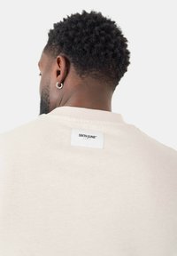 Lys beige bomulds t-shirt med rund hals og et rektangulært mærke på bagsiden med brandnavnet "SIXTH JUNE." Blød tekstur.