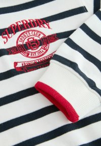Svītraina garām piedurknēm krekls baltā un tumši zilā krāsā ar sarkaniem ribotiem aprocēm. Iezīmēts ar izšūtu logo uzlīmi ar "SUPERDRY" un "SPORTS CLUB".