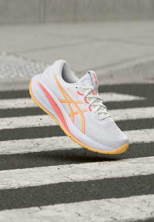 Chaussure de course blanche avec des accents orange et rose, en l'air au-dessus d’un passage piéton rayé noir et blanc dans une rue.