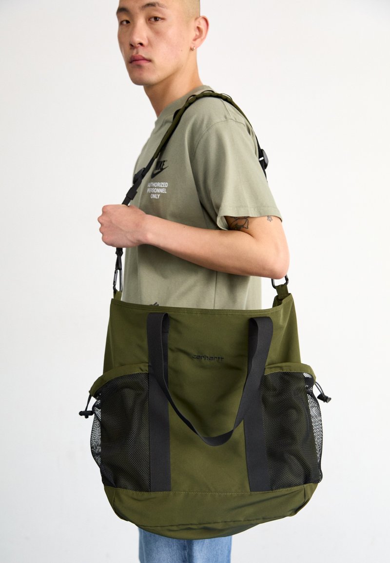 Sac fourre-tout en toile verte avec des poches latérales en filet noir et des anses, arborant un logo Carhartt. Texture lisse, grand avec une forme allongée.