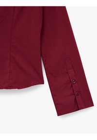Burgundy long-sleeve shirt with a smooth fabric texture. Features a buttoned cuff with two matching buttons and a side slit at the hem.