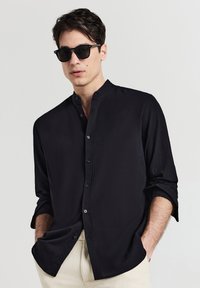 Schwarzes Langarmhemd mit Stehkragen, Knopfverschluss vorne und strukturiertem Stoff. Kombiniert mit einer Sonnenbrille und hellen Hosen.