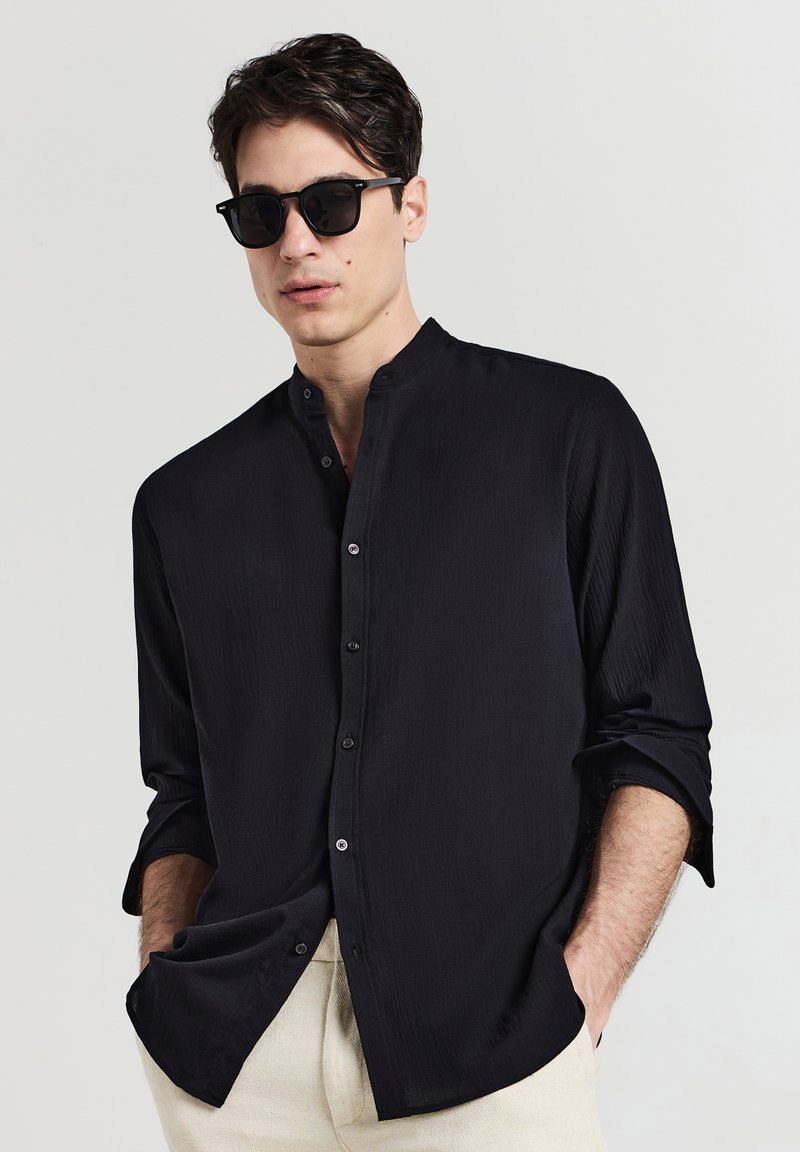 Schwarzes Langarmhemd mit Stehkragen, Knopfverschluss vorne und strukturiertem Stoff. Kombiniert mit einer Sonnenbrille und hellen Hosen.
