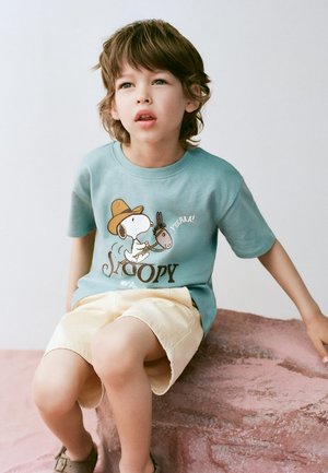 Young child with brown hair wearing a blue Snoopy t-shirt and beige shorts, sitting on a large pink rock, looking slightly upward.