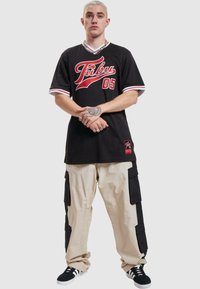 Tricou din plasă neagră cu grafică roșie "Fubu 05", accente albe; pantaloni cargo bej cu buzunare negre; teniși negri cu dungi albe.