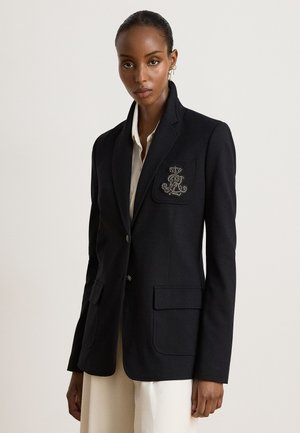 Femme portant un blazer noir avec un blason brodé sur la poche poitrine, une chemise blanche et un pantalon beige sur un fond clair uni.