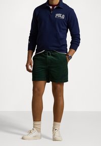 Marinblå zip-up sweatshirt med "POLO"-logotyp, gröna kordshorts, vita sneakers och beiga strumpor. Avslappnad passform med fickor.