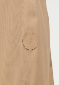 Patch beige rond sur tissu fauve avec canard embossé et texte "SAVE THE DUCK" près d'une couture verticale.