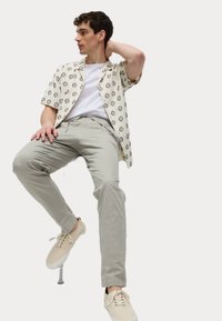 Camisa de manga corta beige con patrón geométrico negro, combinada con pantalones gris claro y zapatillas beige, expuesta sentada en un taburete.