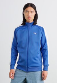 Jachetă de trening albastră cu fermoar, cu accente albe pe umeri, textură netedă și logo-ul Puma pe piept. Are guler înalt și manșete elastice.