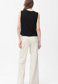 Mujer con top negro sin mangas y pantalones anchos de tiro alto color crema, de pie de espaldas sobre fondo blanco.