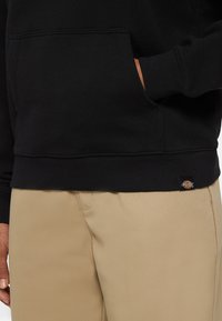 Felpa nera con cappuccio in misto cotone, dotata di tasca frontale e polsini a coste. Indossata con pantaloni kaki che presentano una cucitura sottile. Visibile il marchio Dickies.