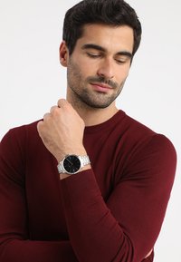 Montre pour homme avec un cadran noir, un bracelet en acier inoxydable argenté, un boîtier rond et un design minimaliste ; portée sur un poignet avec un pull bordeaux.