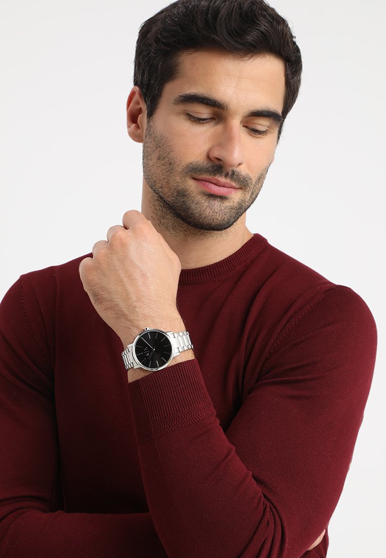 Montre pour homme avec un cadran noir, un bracelet en acier inoxydable argenté, un boîtier rond et un design minimaliste ; portée sur un poignet avec un pull bordeaux.