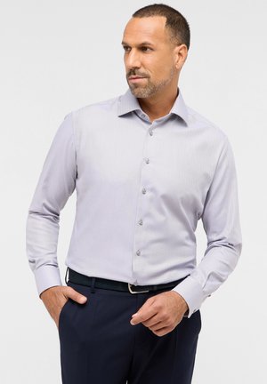 Helle graue Hemdbluse mit dezenten Streifen, langen Ärmeln, spitzem Kragen und knöpfbarem Frontbereich. Kombiniert mit dunklen Hosen und einem Gürtel.