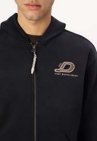 Črna zip-up kapuca z sprednjim žepom, teksturirano blago in bež logotipom s podrobnostmi 'Fort Worth, Texas' na prsih.