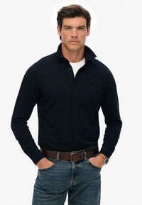 Granatowa koszula z długim rękawem i kołnierzykiem zapinanym na guziki, noszona na białym t-shircie, w połączeniu z niebieskimi dżinsami i brązowym skórzanym paskiem.