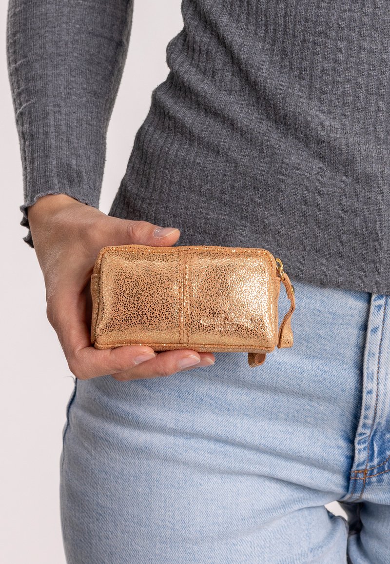 Rose gold rectangular pouch made of textured metallic leather, featuring a zip closure and a small strap, held in a hand against a gray top.