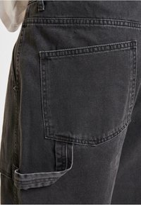 Pantalones de mezclilla negra con un gran bolsillo trasero y un detalle de dobladillo doblado. La tela tiene un acabado texturizado con costuras visibles.