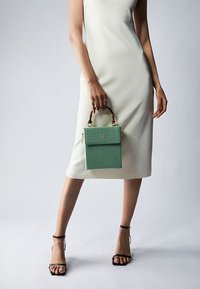 Zielona torebka o fakturze, w prostokątnym kształcie, z materiału sztucznej skóry i górną rączką. Modelka ma na sobie dopasowaną kremową sukienkę i czarne sandały.