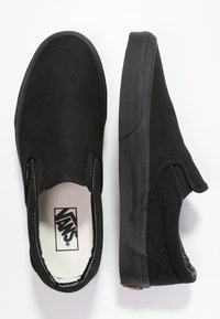 Un paio di Vans nere slip-on è mostrato da una prospettiva dall'alto, mettendo in risalto il tessuto liscio e la suola di gomma. Il logo interno è visibile.