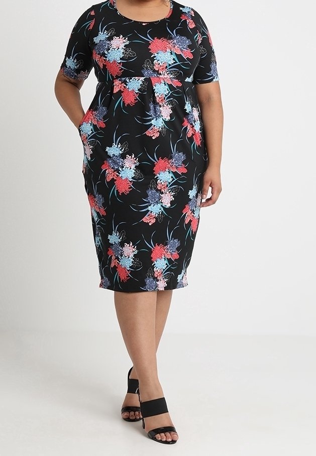 Femme portant une robe noire mi-longue avec un imprimé floral rouge, bleu et rose, manches courtes et talons ouverts noirs.