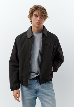 Mladý muž s rozcuchanými vlasmi, oblečený v čiernej bunde so zipsom na sivom tričku a svetlomodrých džínsoch, na jednoduchom bielom pozadí.