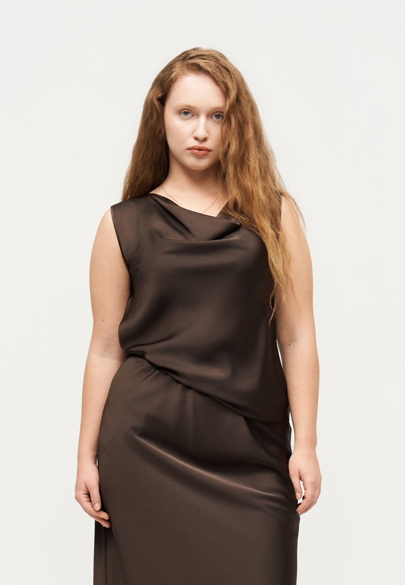 Νεαρή γυναίκα με μακριά κυματιστά κόκκινα μαλλιά, φορώντας ένα αμάνικο σκούρο καφέ σατέν φόρεμα, στέκεται μπροστά σε απλό φωτεινό φόντο.