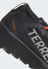 Chaussure de trail noire avec une tige texturée, un logo "TERREX" bien en vue, des accents rouges et une semelle extérieure rugueuse et profilée pour l'adhérence.