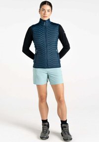 Marineblaue isolierte Weste mit Chevronmuster, Reißverschluss, getragen über einem schwarzen Langarmshirt, kombiniert mit hellblauen Shorts und grauen Wanderschuhen.