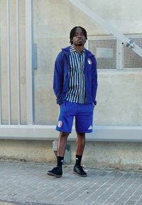Conjunto desportivo azul com blazer de fecho éclair, camisola às riscas e calções. Detalhes incluem uma bandeira italiana e logótipo da Adidas. Sapatos escuros.