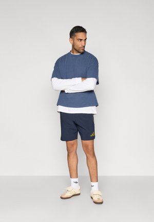 Blauw katoenen T-shirt gedragen over een wit shirt met lange mouwen, gecombineerd met donkerblauwe korte broek met gele strepen en beige sneakers.