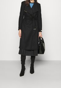 Zwarte wolmix jas met een ceintuur in de taille, grote revers, metallic accenten, gelaagde rok en zwarte suède laarzen tot boven de dijen.