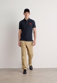 Marineblå polotrøye med rød logo på brystet, khaki bukser og svart caps; modellen har på seg svarte sko og står foran en ensfarget bakgrunn.