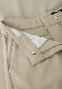 Béžové kalhoty s hladkou texturou mají skrytý zip, zapínání na knoflík a kovovou sponu. Vnitřní kapsa odhaluje bílou podšívku.