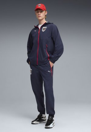 Jonge man draagt een marineblauw sporttrainingspak met rode ritsen en Oostenrijkse voetballogo, een rode pet en zwart-witte Puma sneakers.