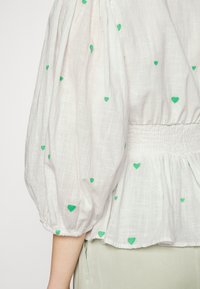 Vit blus med puffärmar, prydd med gröna broderade hjärtformer och en smockad midja. Tillverkad av lätt, texturerad tyg.