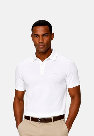 Uomo che indossa una polo bianca aderente infilata in pantaloni beige con cintura marrone, in piedi con le mani in tasca su uno sfondo neutro.