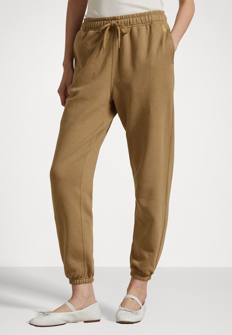 Light brown jogger pants with elastic cuffs and a drawstring waist. Soft, textured fabric; features a small logo on the left thigh.