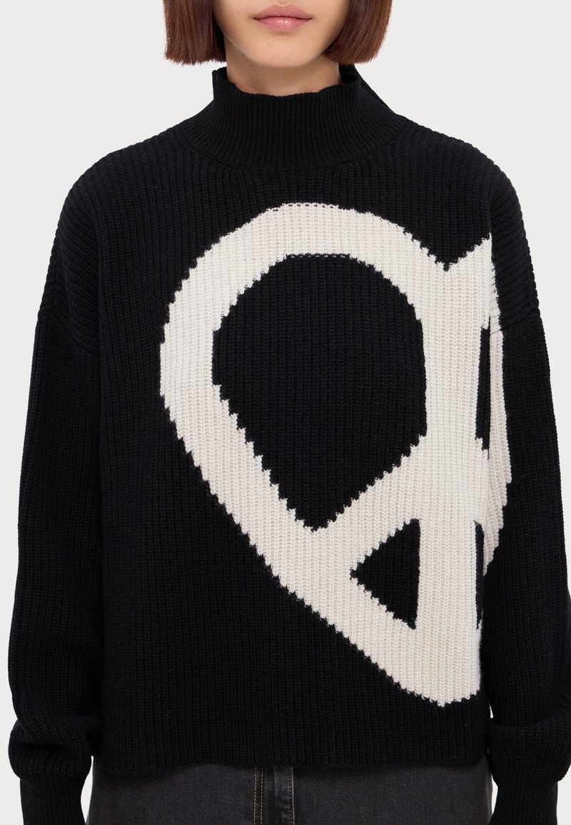 Schwarzer Strickpullover mit einem weißen Friedenssymbols-Design, hoher Ausschnitt und lockere Passform, hervorgehoben durch gerippte Textur.
