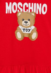 Červená látka s grafikou hnědého medvídka v bílém tričku s nápisem "HRAČKA" nad textem "MOSCHINO" v tučných bílých písmenech.