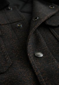 Jachetă din lână texturată, neagră și maro, cu butoane metalice, având două buzunare cu clapetă și guler, evidențiind un model de herringbone.