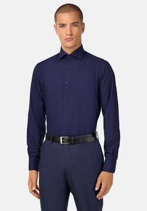 Chemise à manches longues de couleur marine avec un col classique et des boutons sur le devant. Texture lisse et design ajusté. Portée par-dessus un pantalon sombre à motifs et une ceinture noire.