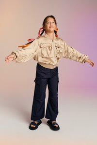 Beige selyem bomber dzseki, két elülső zsebbel, tengerészkék cargo nadrággal és fekete szandállal párosítva. A haj fonatokba van készítve, színes szalagokkal díszítve.