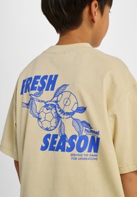 Beige Kurzarm-T-Shirt mit blauem Motiv aus Fußbällen und Blättern sowie dem Schriftzug „FRESH SEASON" und dem „hummel"-Logo.