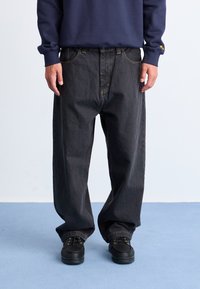 Black denim jeans with a relaxed fit and light yellow stitching. Paired with black shoes, worn with a navy sweatshirt.